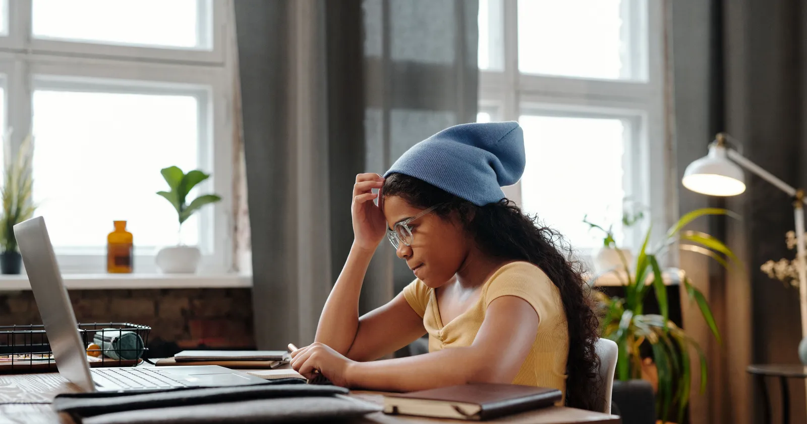 Una niña con gafas y un gorro azul estudia con una computadora portátil en un escritorio junto a una ventana