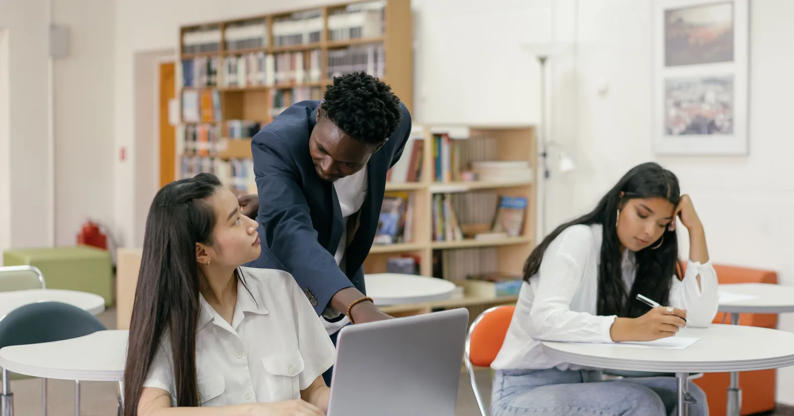 Un profesor se inclina sobre la computadora portátil de un estudiante para ayudarlo durante la clase, mientras otro estudiante escribe en la mesa de al lado