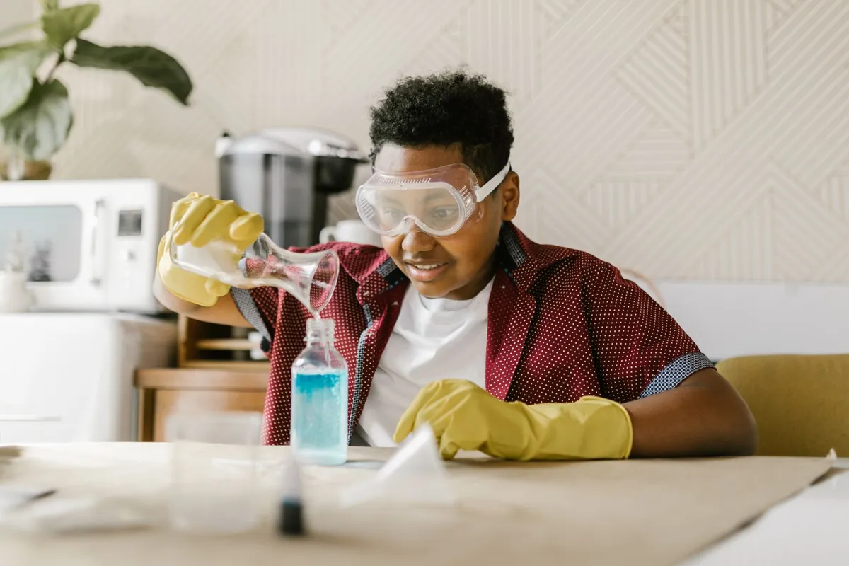 Un niño con gafas de seguridad y guantes de goma vierte un líquido en una botella sobre una mesa común en casa