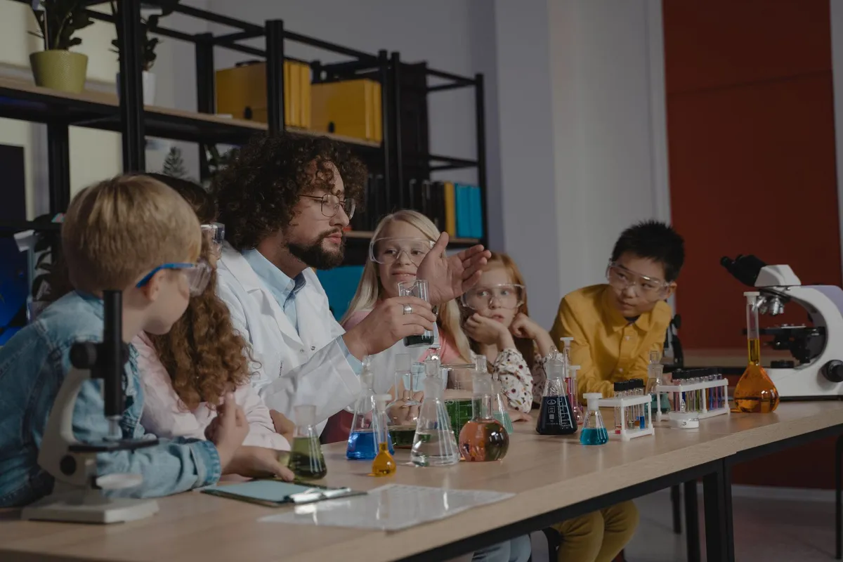 Un profesor con bata de laboratorio hace una demostración de química ante un grupo de estudiantes con gafas de seguridad, alrededor de una mesa llena de matraces