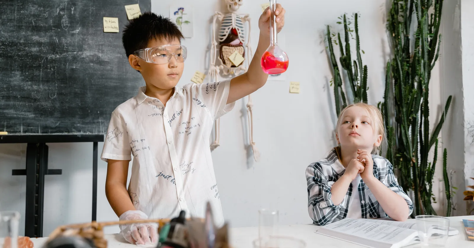 Un estudiante con gafas de seguridad sostiene un matraz con líquido rojo durante un experimento práctico de ciencias, mientras un compañero observa desde la mesa de al lado